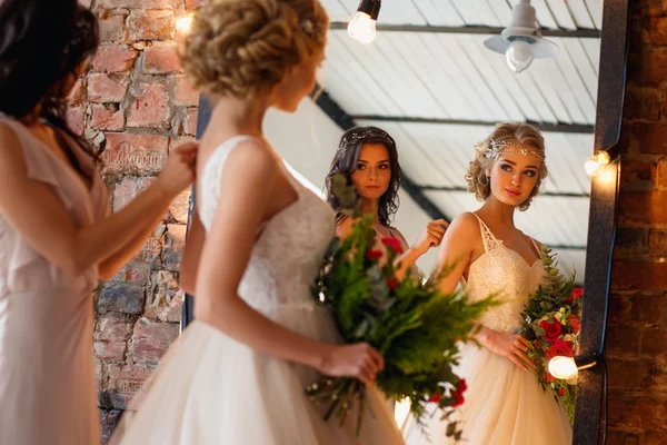 Beautiful blonde bride in luxury wedding dress and pretty bridesmaid in a morning in a loft space with a mirror and garland of lamps. Fashion modern wedding photo.