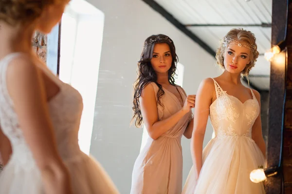 Beautiful blonde bride in luxury wedding dress and pretty bridesmaid in a morning in a loft space with a mirror and garland of lamps. Fashion modern wedding photo.
