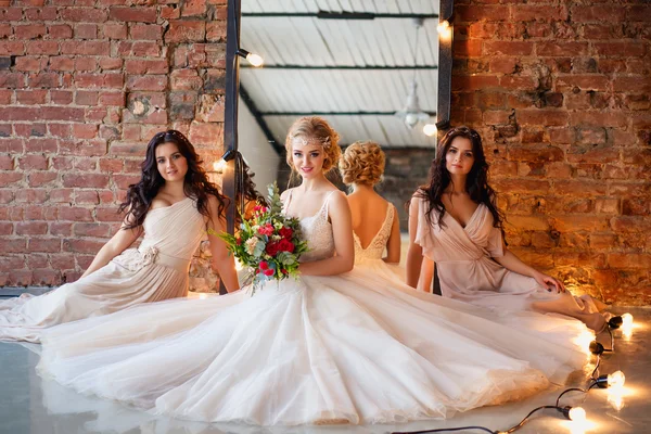 Beautiful blonde bride in luxury wedding dress and pretty twins bridesmaids in similar dresses in a morning in a loft space with a mirror and garland of lamps. Fashion modern wedding photo.
