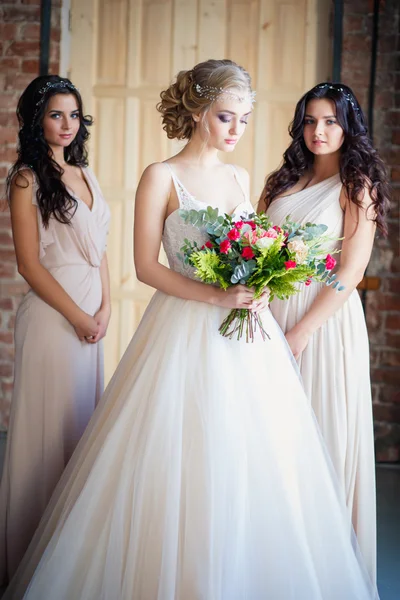 Beautiful blonde bride in luxury wedding dress and pretty twins bridesmaids in similar dresses in a morning in a loft space. Fashion modern wedding photo