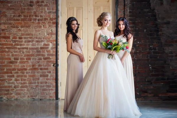 Beautiful blonde bride in luxury wedding dress and pretty twins bridesmaids in similar dresses in a morning in a loft space. Fashion modern wedding photo