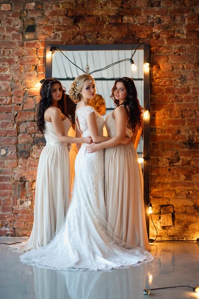 Beautiful blonde bride in luxury wedding dress and pretty twins bridesmaids in similar dresses in a morning in a loft space with a mirror and garland of lamps. Fashion modern wedding photo.