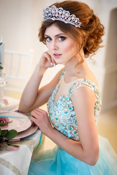 Beautiful young lady in a luxury blue dress sitting by a table with a bouquet of flowers