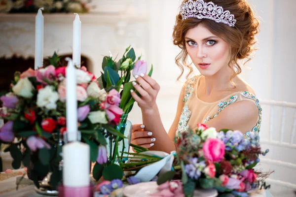 Beautiful young lady in a luxury blue dress sitting by a table with a bouquet of flowers