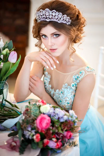 Beautiful young lady in a luxury blue dress sitting by a table with a bouquet of flowers