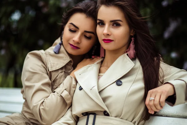 Two beautiful twins young women in trench coats near blooming lilac