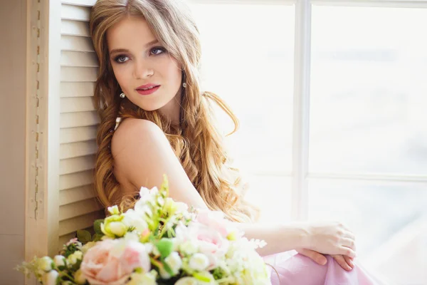 Portrait of pretty young girl in a sweet dress and long blonde hair sitting on a window with flowers composition