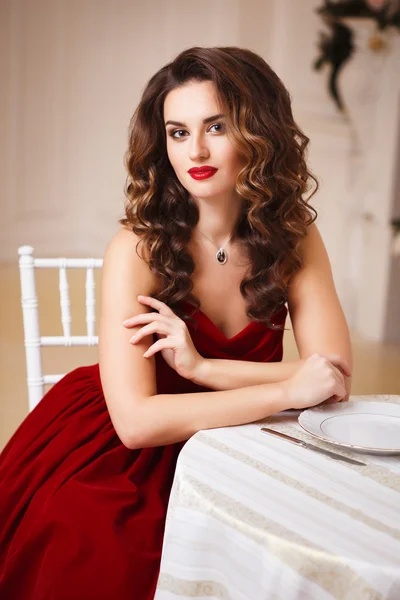 Close-up portrait of beautiful young woman in gorgeous red velvet evening dress sitting by the table in expensive interior