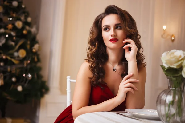 Close-up portrait of beautiful young woman in gorgeous red velvet evening dress sitting by the table in expensive interior