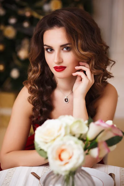 Close-up portrait of beautiful young woman in gorgeous red velvet evening dress sitting by the table in expensive interior