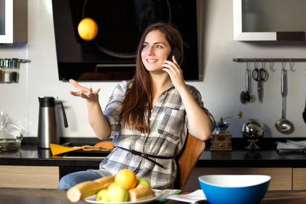 Portrait of young brunette woman in a kitchen talking by mobile phone and toss orange