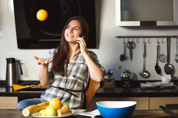 Portrait of young brunette woman in a kitchen talking by mobile phone and toss orange