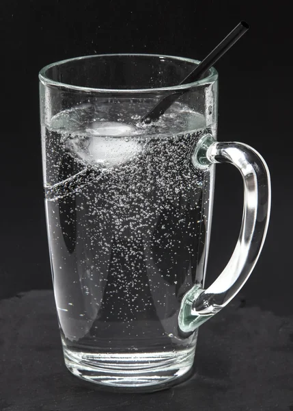 Glass refreshing drink, soda water with ice in a glass with ice cubes and cocktail drinking straw in black background, against thirst on a hot day. Water is the main source of everyday life