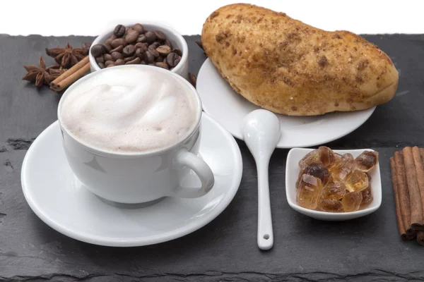 Cup of coffee cappuccino with milk foam on a saucer, fresh baked bun, caramelized sugar, coffee beans, cinnamon. Popular morning Breakfast. on a black background plates of slate