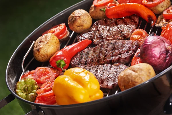 Assorted delicious grilled meat steak with various vegetables, paprika, tomato, pepper, potato, over the coals on a round modern black barbecue close up - weekend or vacation concept