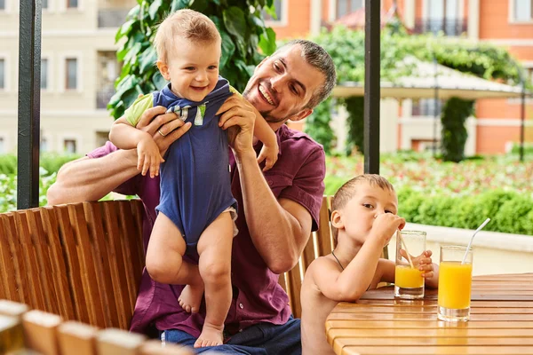 Smiling Father holding happy baby and another boy drinking orange juice
