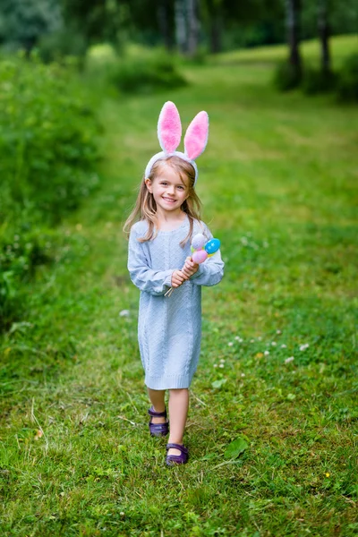 Adorable little girl with long blond hair wearing rabbit or bunny ears and blue knitted dress. Smiling little girl holding bunch of painted colour eggs and walking in spring park. Easter celebrations