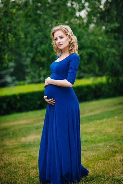 Pretty young pregnant woman in blue dress with long blond curly hair holding her belly and looking at camera in summer park on rainy day. Pregnancy and femininity concept. Waterdrops on blue dress