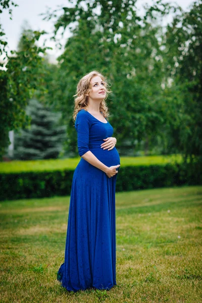 Pretty young pregnant woman in blue dress with long blond curly hair holding her belly, smiling and looking at sky in summer park on rainy day. Pregnancy and femininity concept. Waterdrops on dress