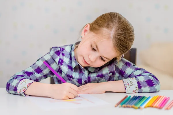 Beautiful pensive little girl with blond hair sitting at white table and drawing with multicolored pencils