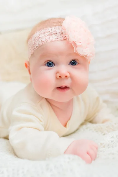 Amazed baby girl with chubby cheeks and big blue eyes wearing white clothes and pink band with flower lying on bed. Babyhood and childhood concept