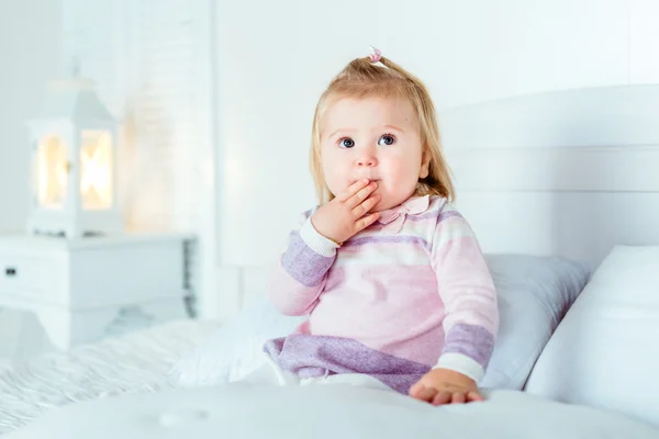 Funny surprised blond little girl sitting on bed in bedroom. White interior, big bed, bedside table and night lamp. Girl touches her mouth by hand