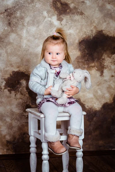 Displeased blond little girl sitting on white chair and holding her toys. Studio portrait on brown grunge background