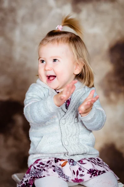 Cute little girl with blond hair sitting on chair, laughing and clapping her hands. Studio portrait on brown grunge background
