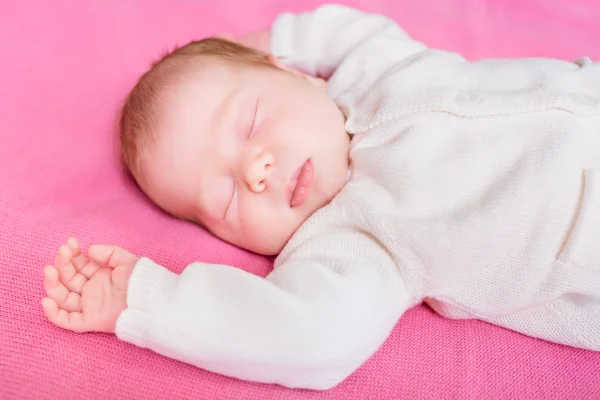 Cute little baby with closed eyes wearing knitted white clothes lying on pink plaid. 2 week old baby sleeping on pink sofa. Security and childcare concept