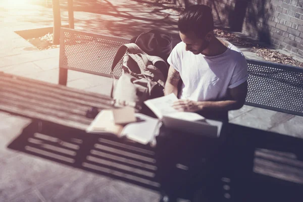 Young man wearing white tshirt sitting city park and reading book. Studying at the University, preparation for exams. Books, notebook, backpack bench. Horizontal, blurred, film effect