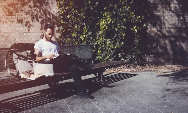 Young man wearing white tshirt sitting city park and reading book. Studying at the University, working new project. Books, laptop, backpack bench. Horizontal,  film effect