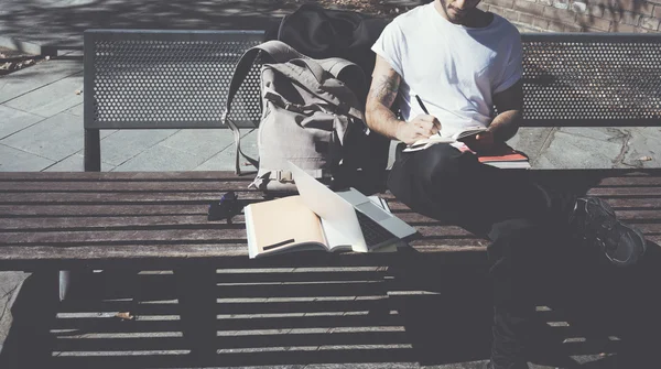 Hipster wearing white tshirt sitting city park bench and writing textbook. Studying at the University, working project. Books, generic design laptop, backpack bench. Horizontal, blurred, film effect
