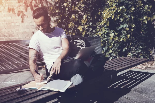 Student wearing white tshirt sitting city park bench and writing textbook message. Studying at the University, preparation for exams. Books, generic design laptop, backpack bench. Horizontal