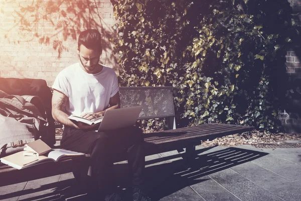 Stylish man wearing white tshirt sitting city park and reading book. Studying at the University, working new project,  preparation for exams. Books, laptop, backpack bench. Horizontal