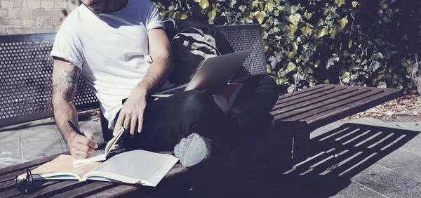 Man wearing white tshirt sitting city park and writes message. Studying at the University, working project. Books, generic design laptop, backpack bench. Wide