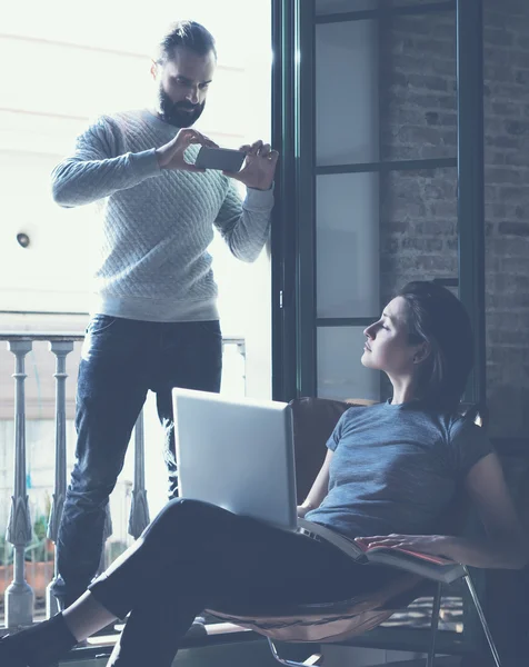 Young couple work together. Bearded man making photo smartphone beautiful girl. Working with new startup project in modern loft. Using contemporary notebook. Vertical, film effect.