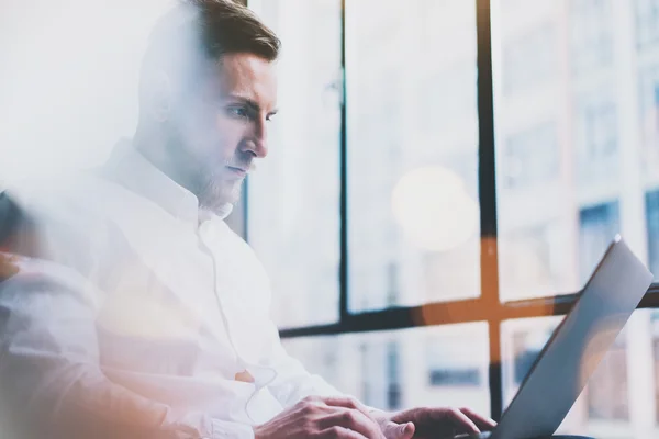 Bearded young businessman working on modern loft office. Man wearing white shirt and using contemporary laptop. Panoramic windows background. Horizontal, film effect, bokeh