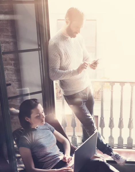 Young couple work together. Photo woman and bearded man working with new startup project in modern loft. Using contemporary notebook, smartphone. Vertical, film effect. Blurred background