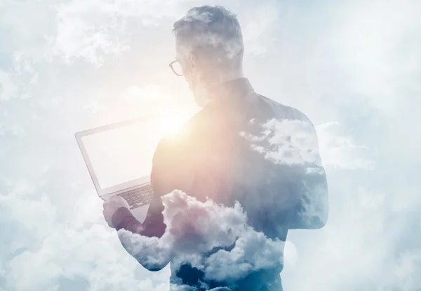 Double exposure clouds young bearded businessman wearing black shirt and holding contemporary notebook hands. White empty screen ready for you message. Sunlight effect. Horizontal mockup