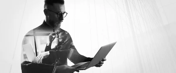 Double exposure bearded businessman wearing shirt and glasses,holding contemporary notebook hands. Portrait young banker using laptop in modern office, skyscraper background. Wide, black,white
