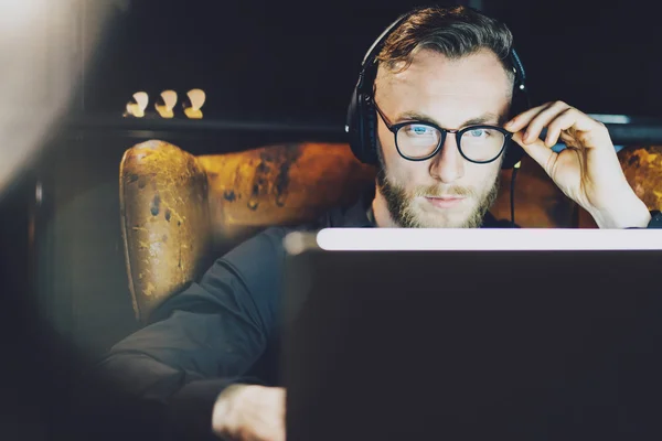 Photo stylish bearded man wearing glasses working modern loft office. Young banker sitting vintage chair,listening music laptop night.Using contemporary notebook.Horizontal,film effect