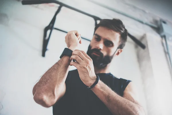 Young Bearded Sportive Man Workout Session Checks Fitness Results Smart Watch.Adult Guy Dress Sport Tracker Wristband Arm.Training hard gym.Horizontal bar background.Blurred.