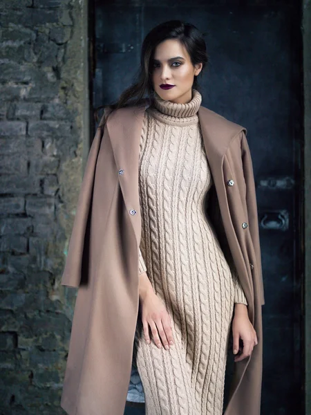 Elegant fashion model wearing long mocha coat, posing before a door.