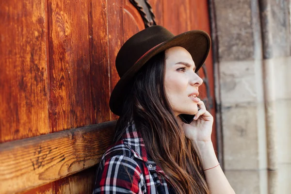 Young woman with a hat next to an old wooden door talking at cel