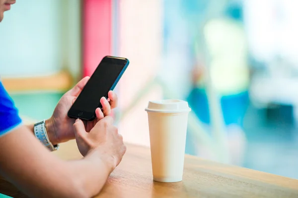 Closeup of male hands holding cellphone and class of cofee in cafe. Man using mobile smartphone. Boy touching a screen of his smarthone. Blurred background, horizontal.