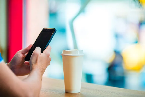 Closeup of male hands holding cellphone and glass of coffee in cafe. Man using mobile smartphone. Boy touching a screen of his smarthone. Blurred background, horizontal.