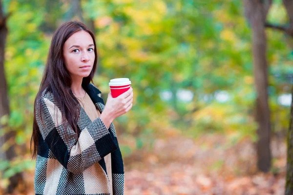 Beautiful woman drinking coffee in autumn park under fall foliage. Coffee to go in her hands