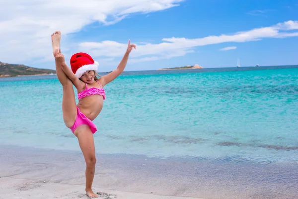 Smiling little girl making stretching exercise in Santa Hat on white beach