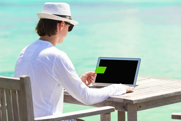 Young man working on laptop with credit card during tropical vacation