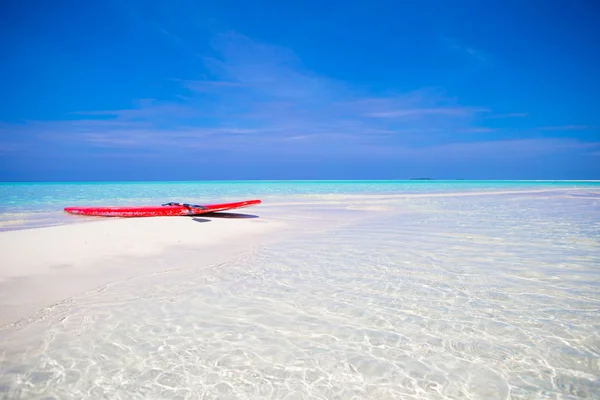 Red surfboard on white sandy beach with turquoise water at tropical island in Indian Ocean